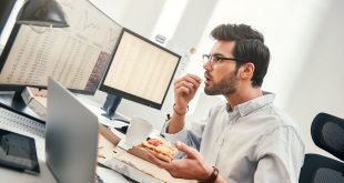 Man sitting at bank of computers eating pizza and looking stressed.