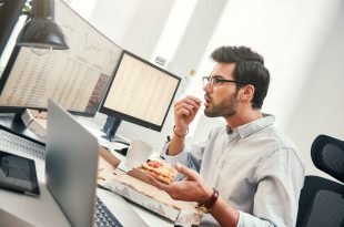 Man sitting at bank of computers eating pizza and looking stressed.