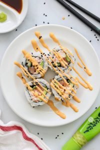 Spicy Salmon Roll cut up on a white plate with spicy mayo drizzled on top and chopsticks and wasabi nearby.