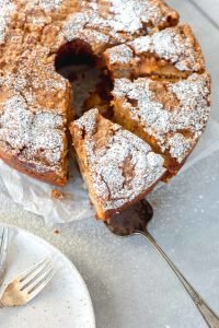 A picture of a Jewish apple cake. It is shaped like an angel food cake and has one slice cut and partially removed. The cake has powdered sugar on top.