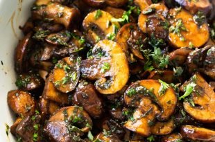 a close up photo of golden sauteed mushrooms topped with fresh oregano and thyme in a large white serving bowl.